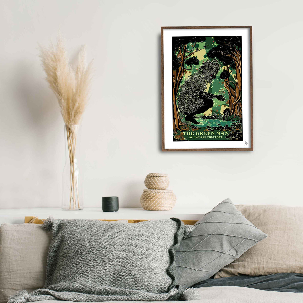 A framed art print titled 'The Green Man' featuring a vintage-style forest illustration, displayed on a white wall above a modern white sideboard with neutral-toned pillows and a vase of dried grass.