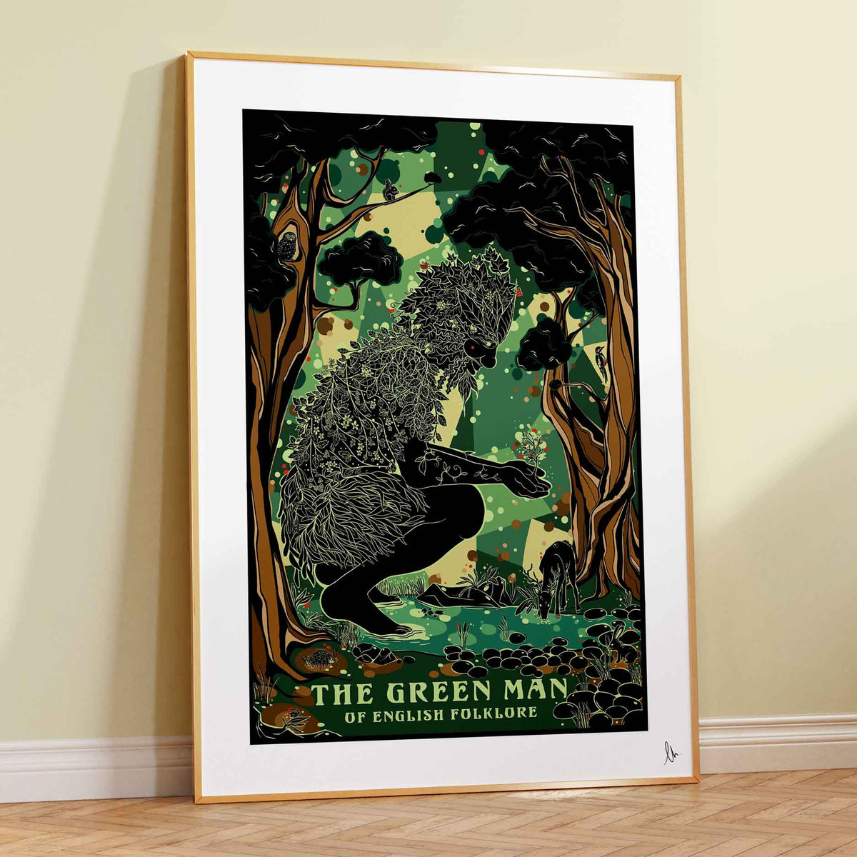 A framed art print of 'The Green Man of English Folklore' leaning against a cream-colored wall on a light wood herringbone floor. The poster features a black silhouette of a figure made of leaves and branches, set against a lush green forest background, enclosed in a white mat and a light wood frame.