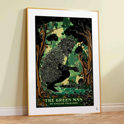 A framed art print of 'The Green Man of English Folklore' leaning against a cream-colored wall on a light wood herringbone floor. The poster features a black silhouette of a figure made of leaves and branches, set against a lush green forest background, enclosed in a white mat and a light wood frame.