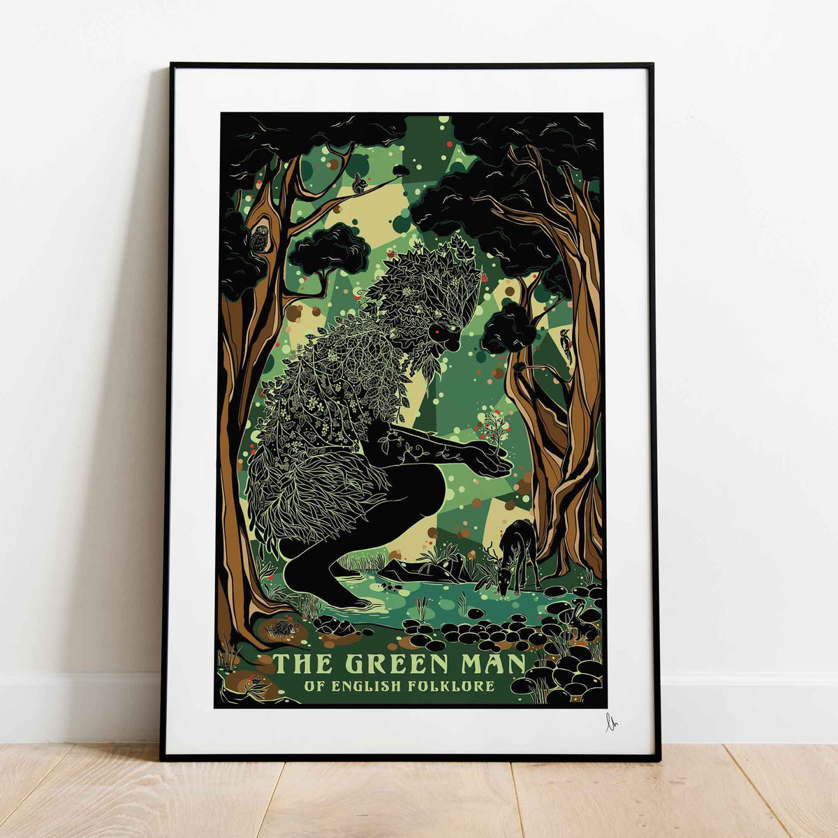 A framed art print of 'The Green Man' from English Folklore leaning against a white wall on a light wood floor. The print features a black and green illustration of a figure in a forest with the title text at the bottom.