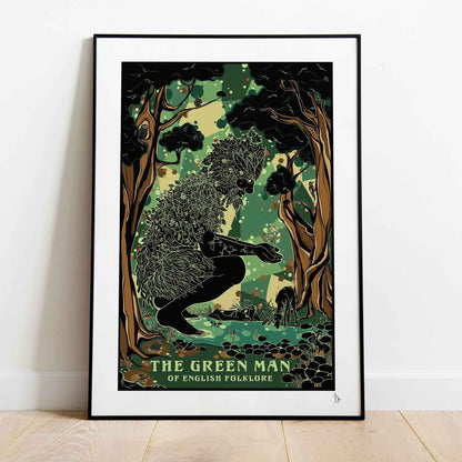 A framed art print of 'The Green Man' from English Folklore leaning against a white wall on a light wood floor. The print features a black and green illustration of a figure in a forest with the title text at the bottom.
