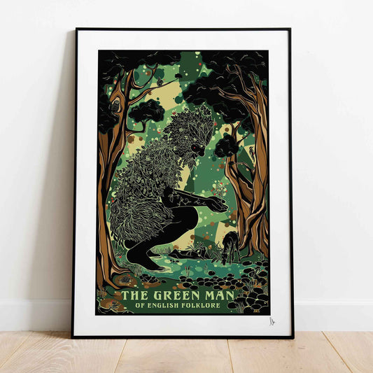 A framed art print of 'The Green Man' from English Folklore leaning against a white wall on a light wood floor. The print features a black and green illustration of a figure in a forest with the title text at the bottom.